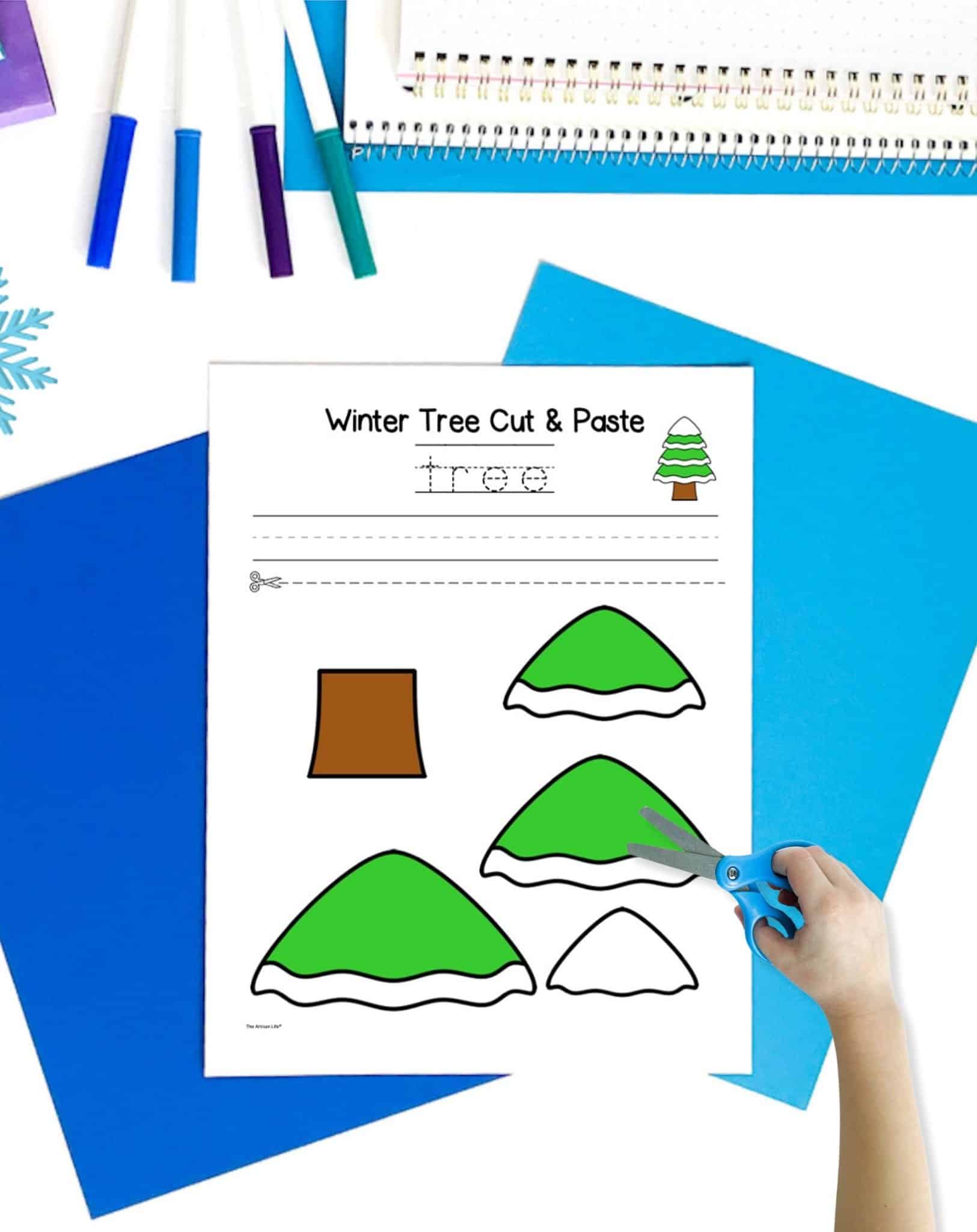 a colored winter tree craft printable on a table with blue papers. A child's hand holds scissors in the foreground