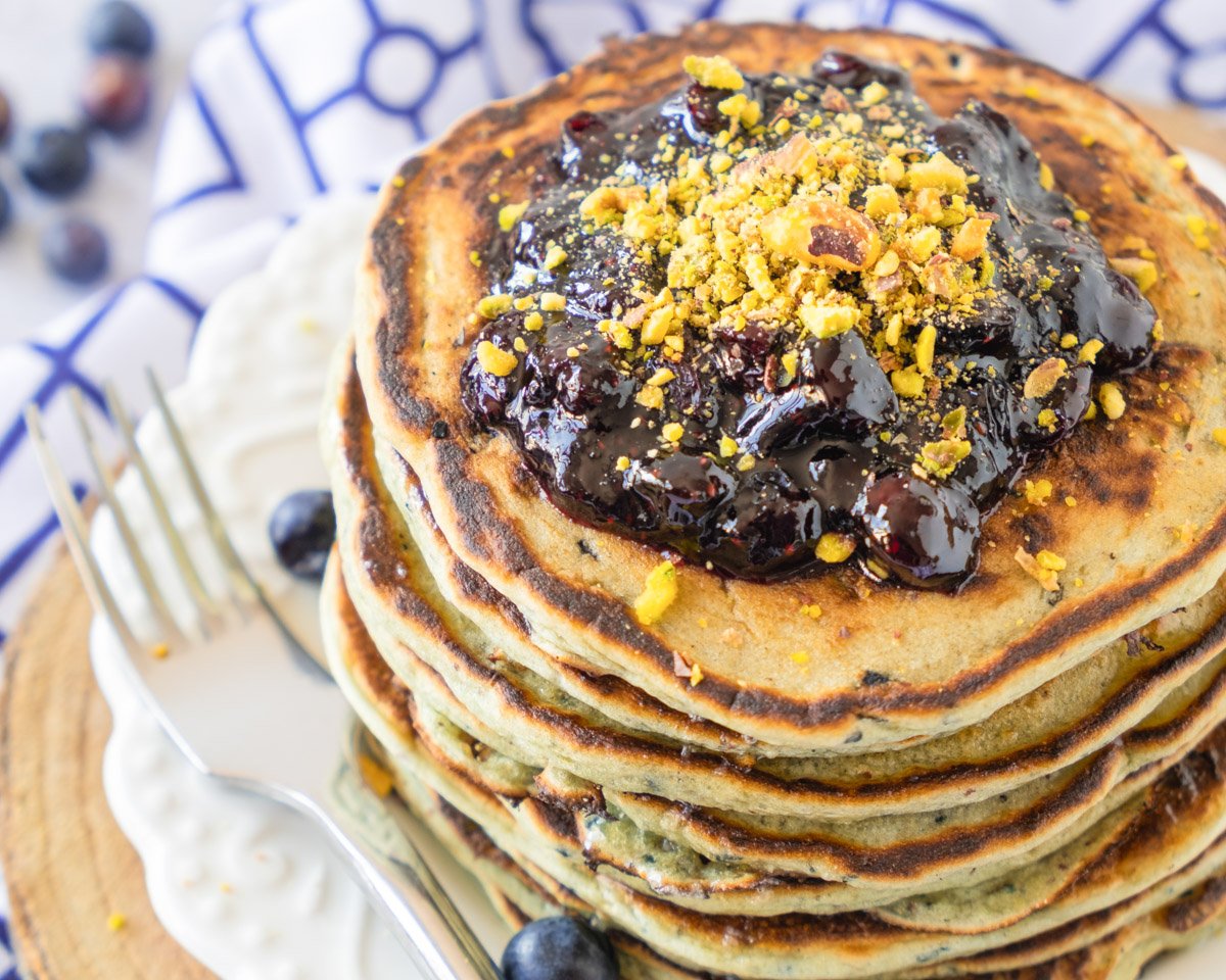 A stack of gluten-free blueberry pancakes topped with blueberry sauce and garnished with crushed nuts is displayed on a white plate, emphasizing a homemade breakfast option.
