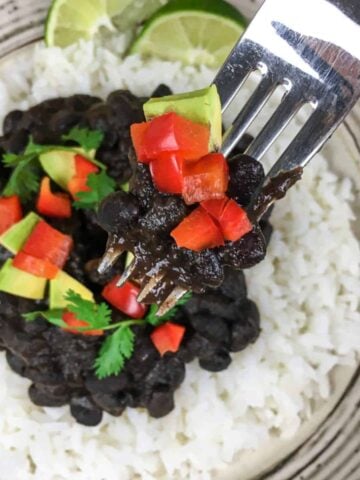 a plate with white rice and black beans. A fork with black beans, red pepper, and avocado is lifted towards the camera.