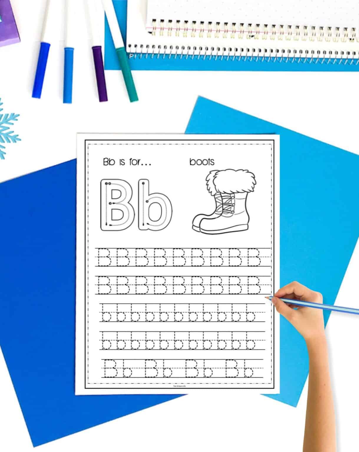 a letter b tracing worksheet with winter boots. The page is on a. table with blue papers. A child's hand holding a pencil is visible in the foreground