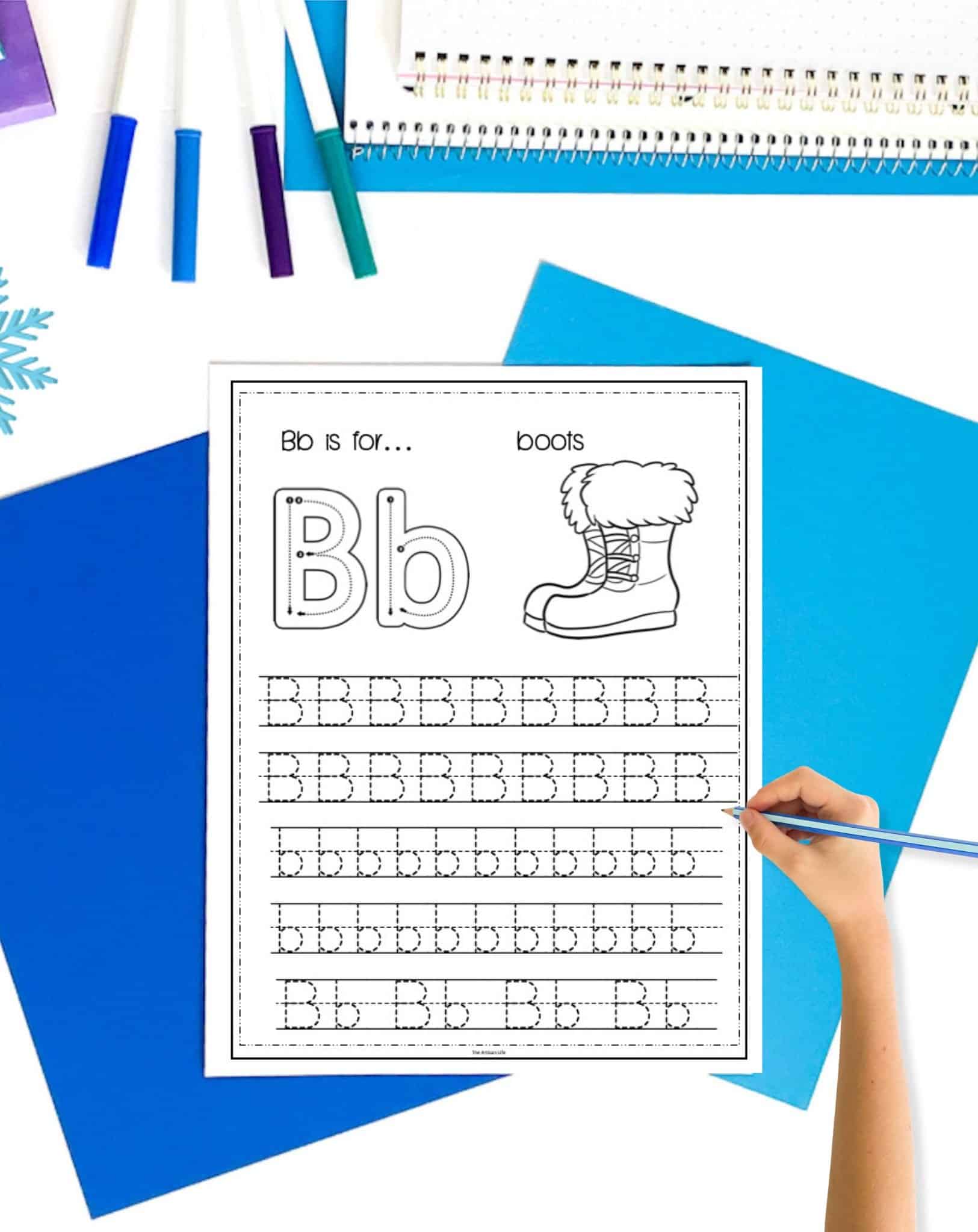 a letter b tracing worksheet with winter boots. The page is on a. table with blue papers. A child's hand holding a pencil is visible in the foreground 