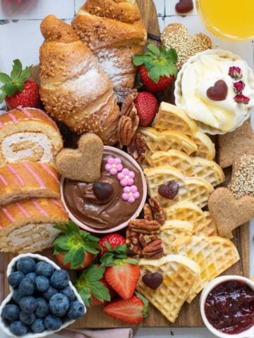 a top down photo of a breakfast board with heart shaped waffles, fruit and spreads like cream cheese and jelly