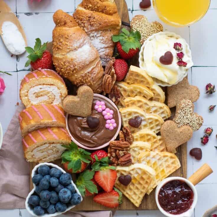 a top down photo of a breakfast board with heart shaped waffles, fruit and spreads like cream cheese and jelly