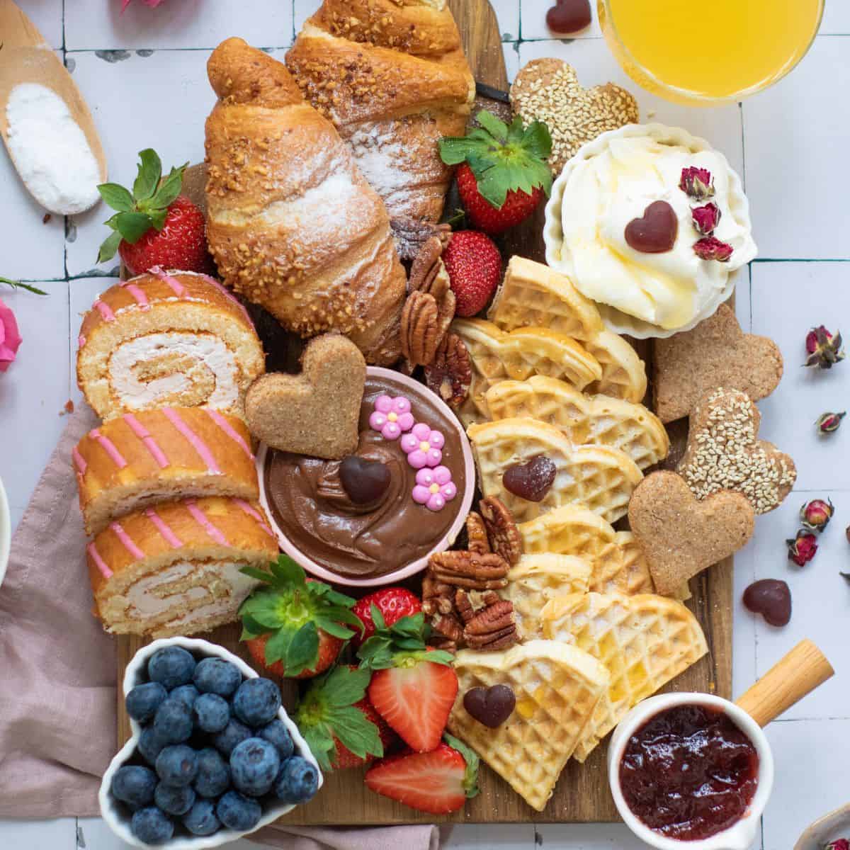 a top down photo of a breakfast board with heart shaped waffles, fruit and spreads like cream cheese and jelly