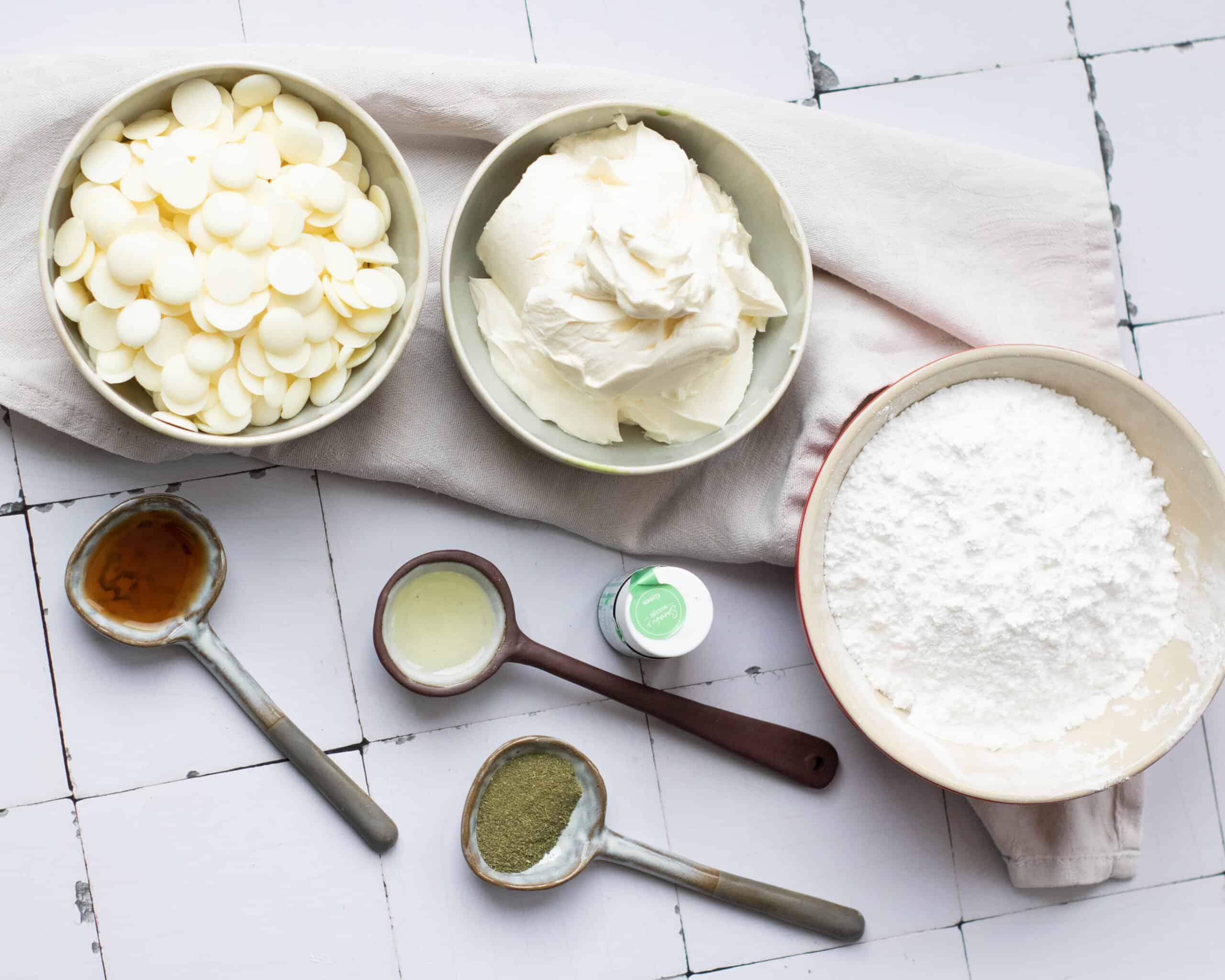 a top down image of bowls with lowered sugar, white chocolate, and cream cheese on a tile counter with matcha powder, coconut oil, vanilla extract, and green gel food coloring 