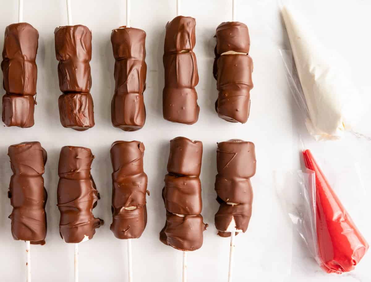 chocolate covered marshmallows on lollipop sticks next to icing bags with red and white chocolate 