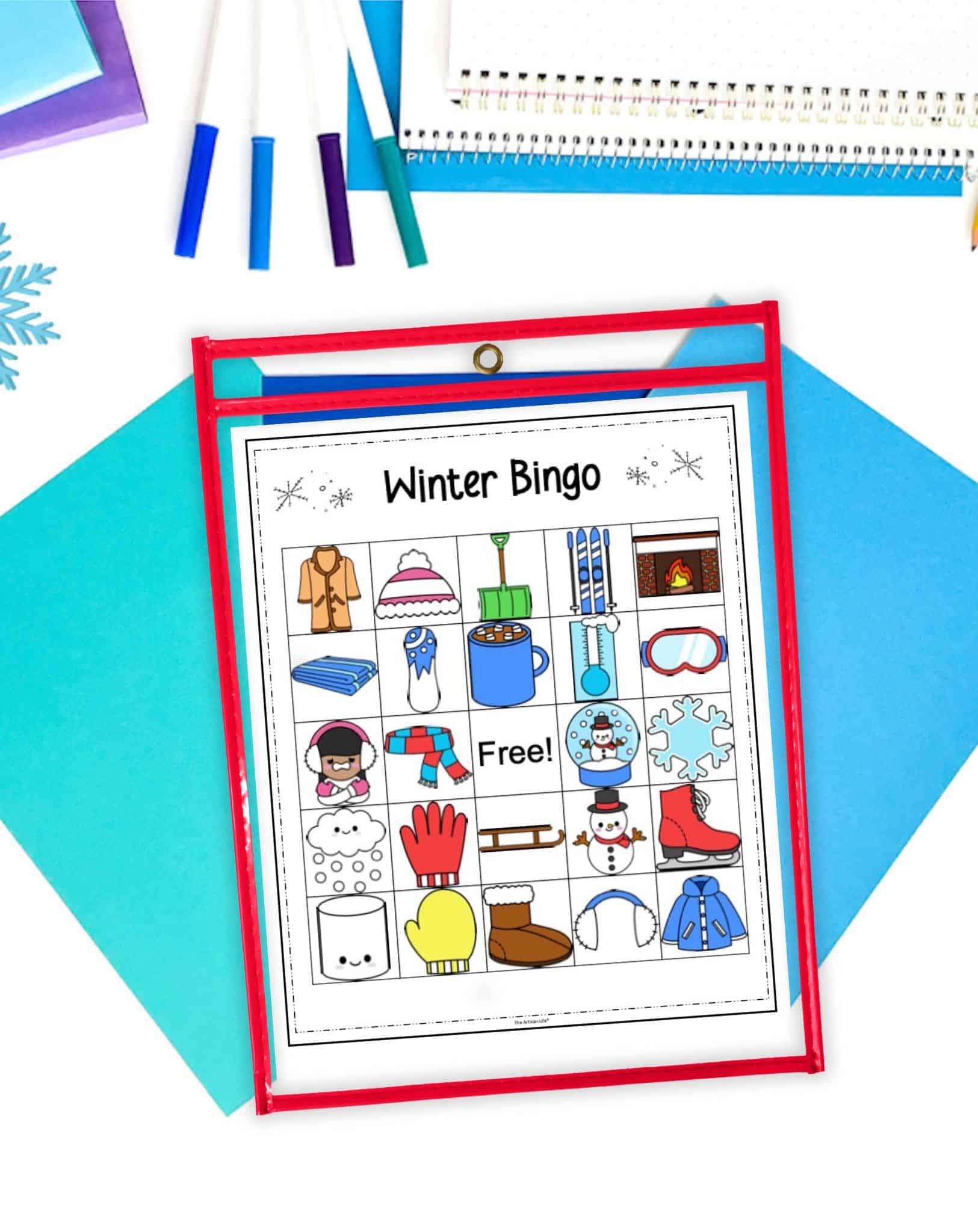 a winter picture bingo card for kids in a dry erase pocket. The pocket is on a desk with blue papers and markers