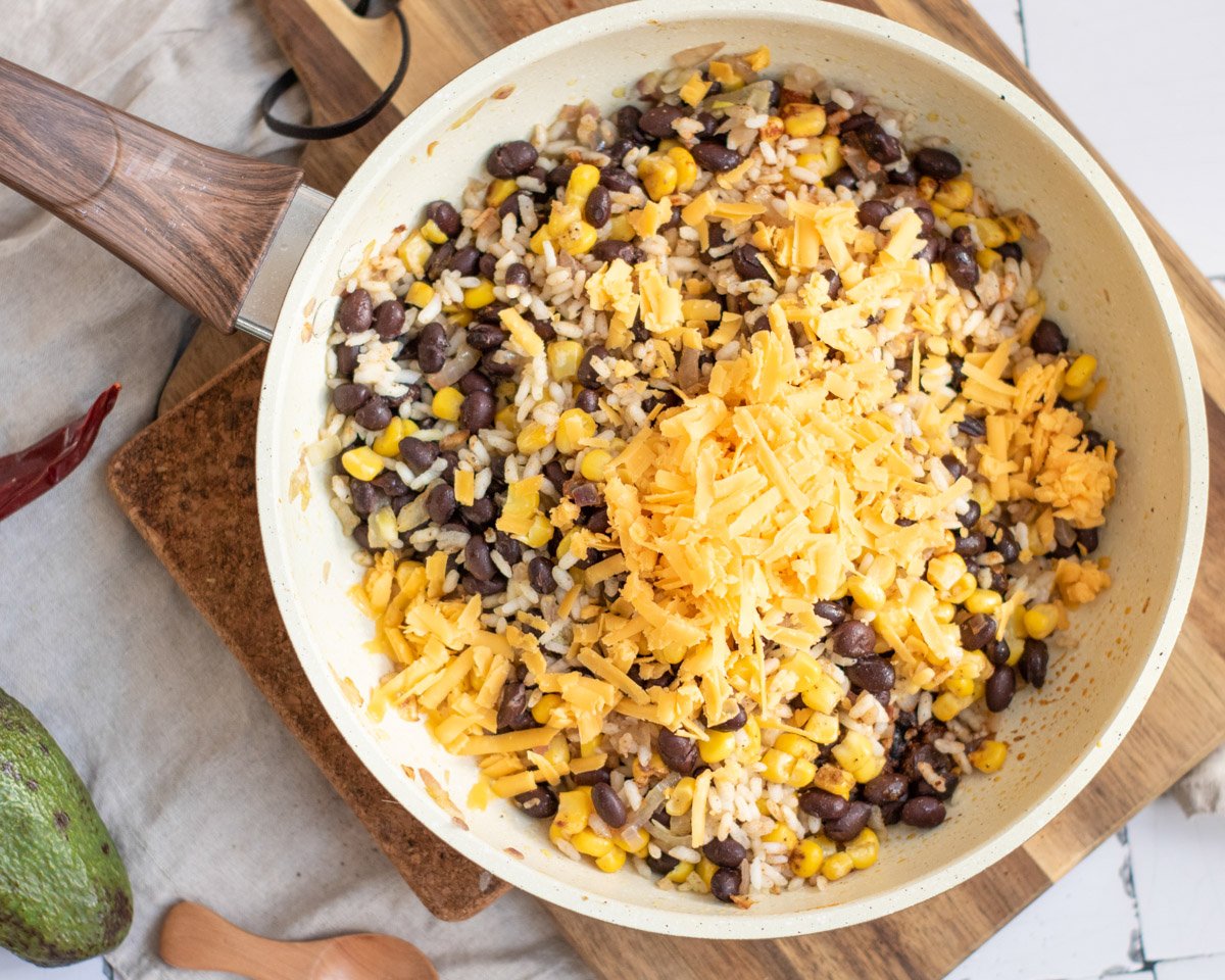 A skillet filled with a mixture of black beans, corn, and rice topped with shredded cheese, prepared as a filling for vegetarian enchiladas.
