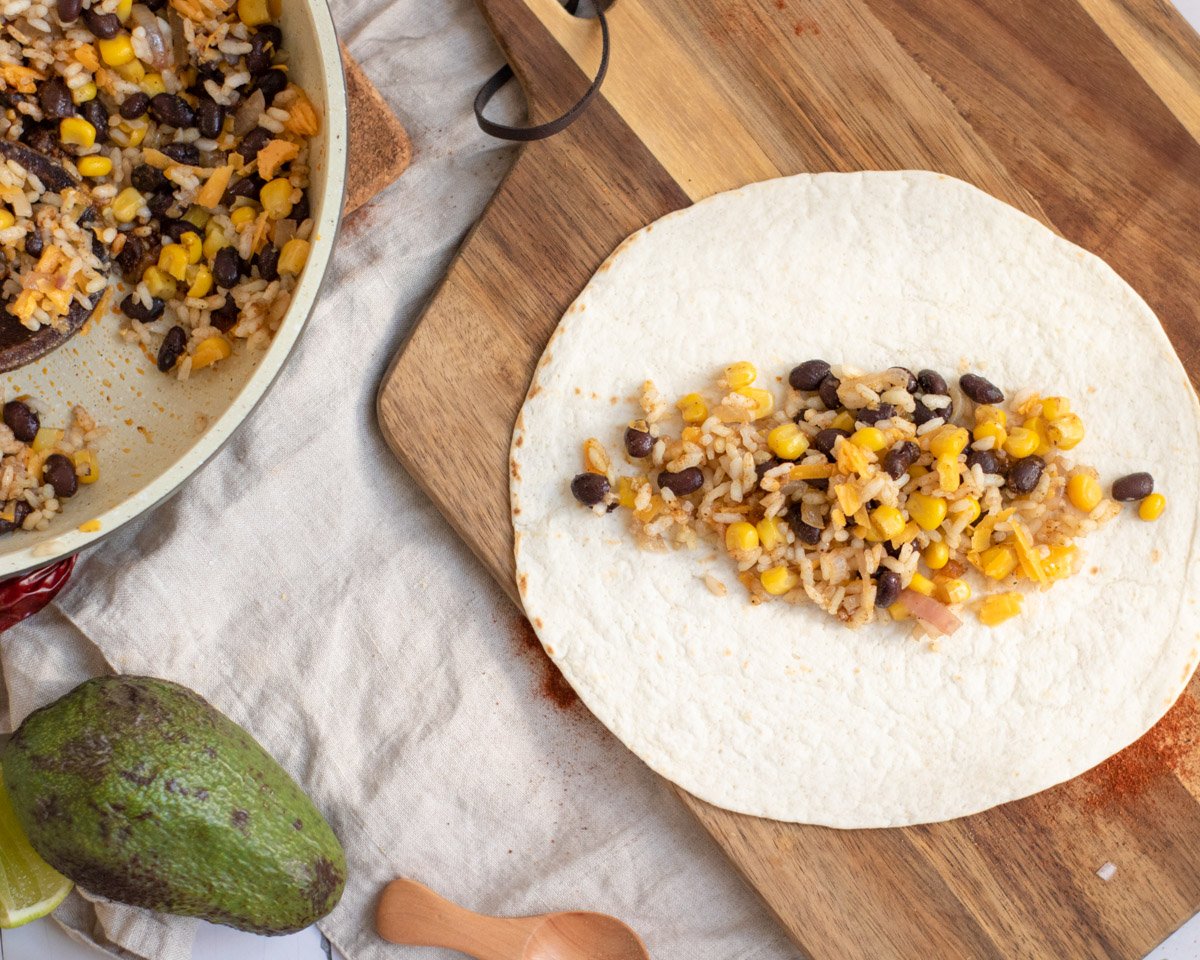 A tortilla is filled with a mixture of rice, black beans, and corn, accompanied by an avocado and lime on a textured surface, suggesting preparation for healthy vegetarian enchiladas.
