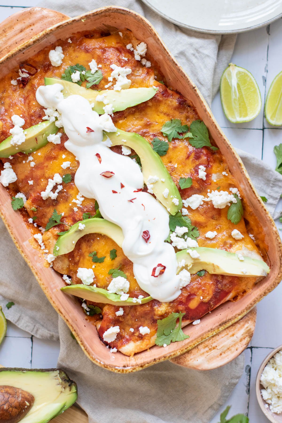 A baked dish of black bean enchiladas topped with melted cheese and served with lime wedges and a side of cheese, placed on a beige cloth.
