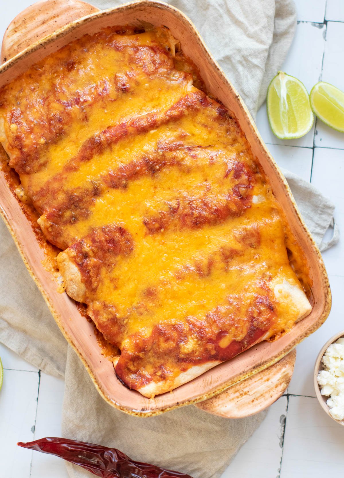 A rectangular baking dish contains black bean enchiladas topped with shredded cheese and red sauce, placed on a beige cloth with an avocado and a dried pepper nearby.
