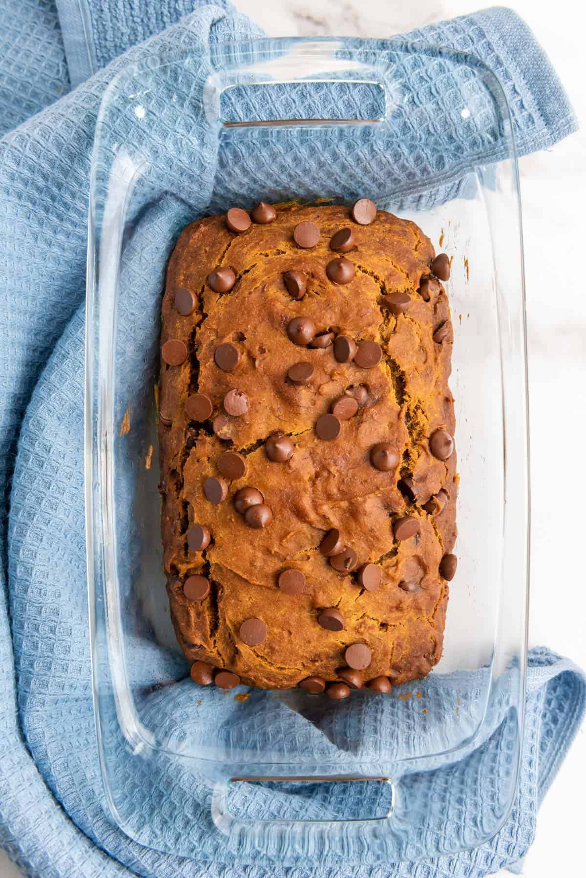 A loaf of gluten-free banana bread with chocolate chips is placed in a glass dish on a blue cloth.
