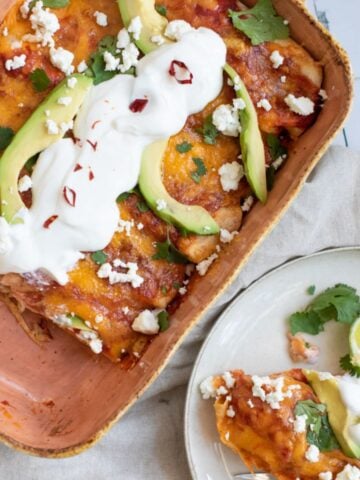 A casserole dish holds black bean enchiladas topped with avocado, sour cream, and cilantro, served alongside a plate with a portion of the enchiladas and lime wedges.