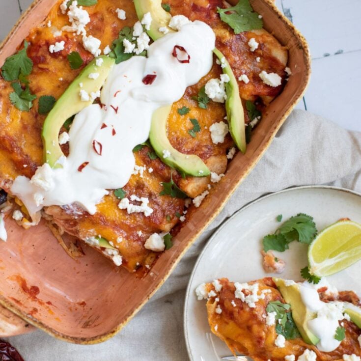 A casserole dish holds black bean enchiladas topped with avocado, sour cream, and cilantro, served alongside a plate with a portion of the enchiladas and lime wedges.