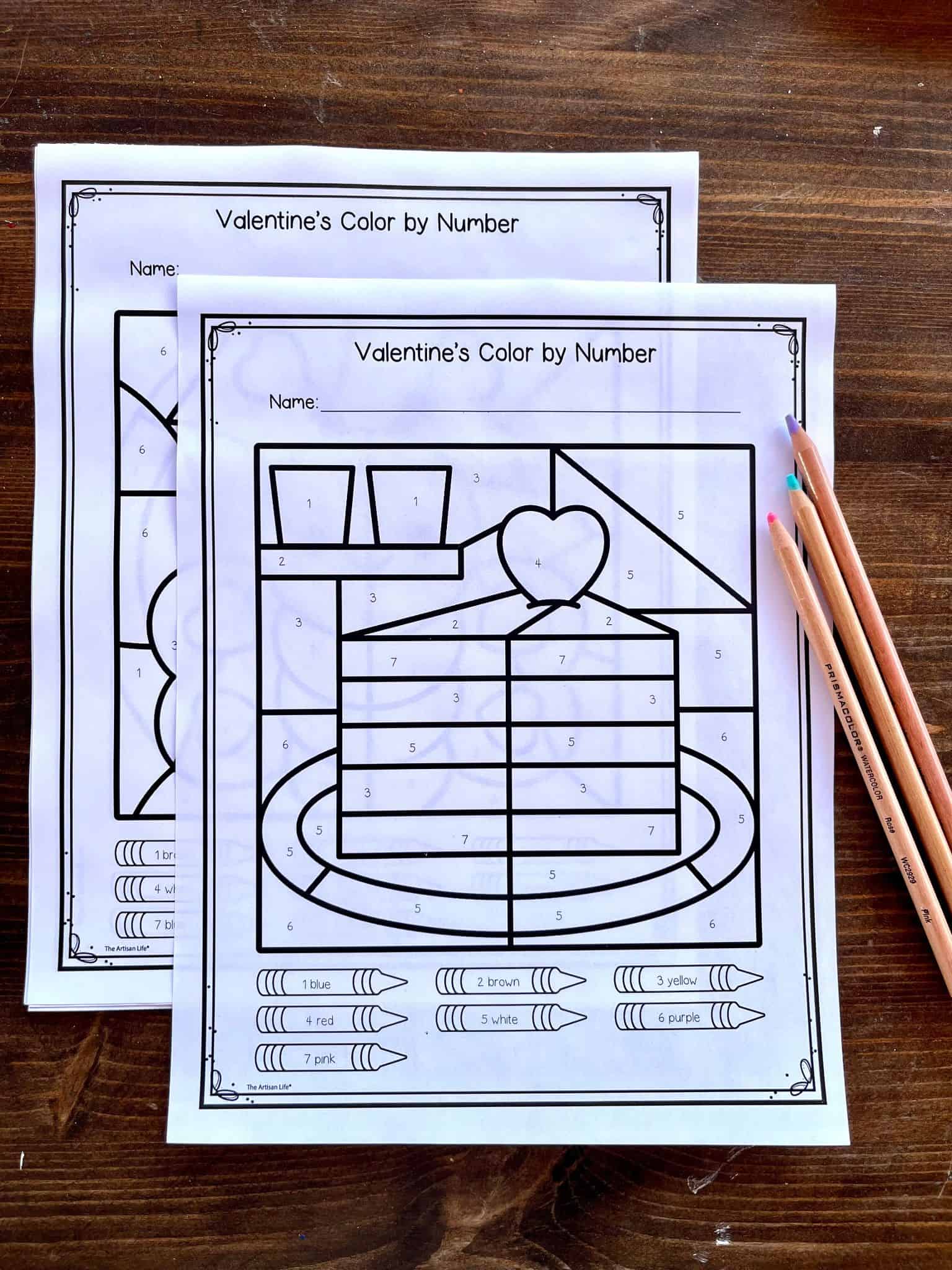 Two printed "Valentine's Color by Number" worksheets are placed on a wooden surface alongside colored pencils, featuring box outlines and a heart design for kids' number recognition and coloring activities related to Valentine’s Day.
