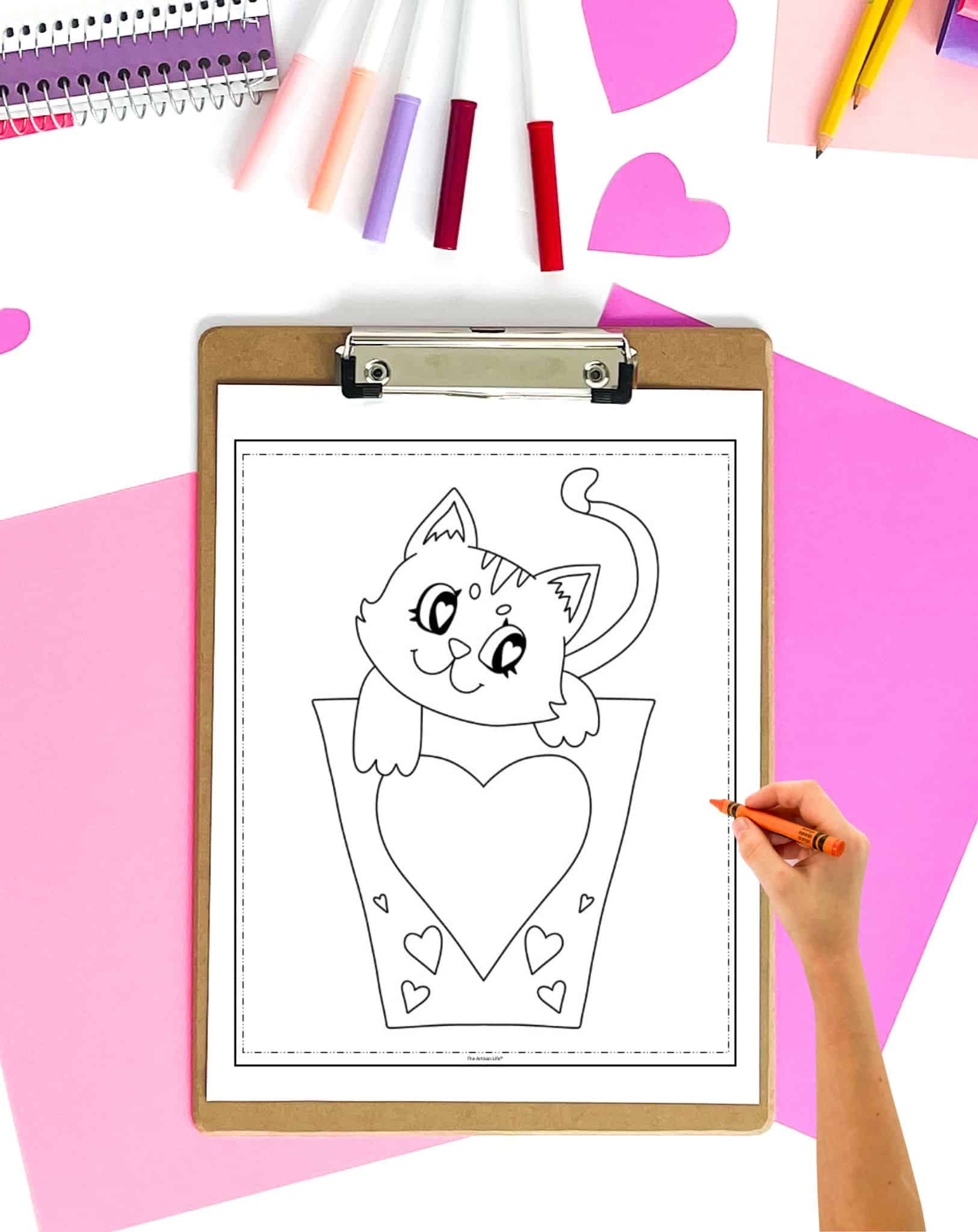 A hand is coloring a printable Valentine's Day coloring page featuring a cat in a heart-shaped container, surrounded by various craft supplies and heart cutouts.
