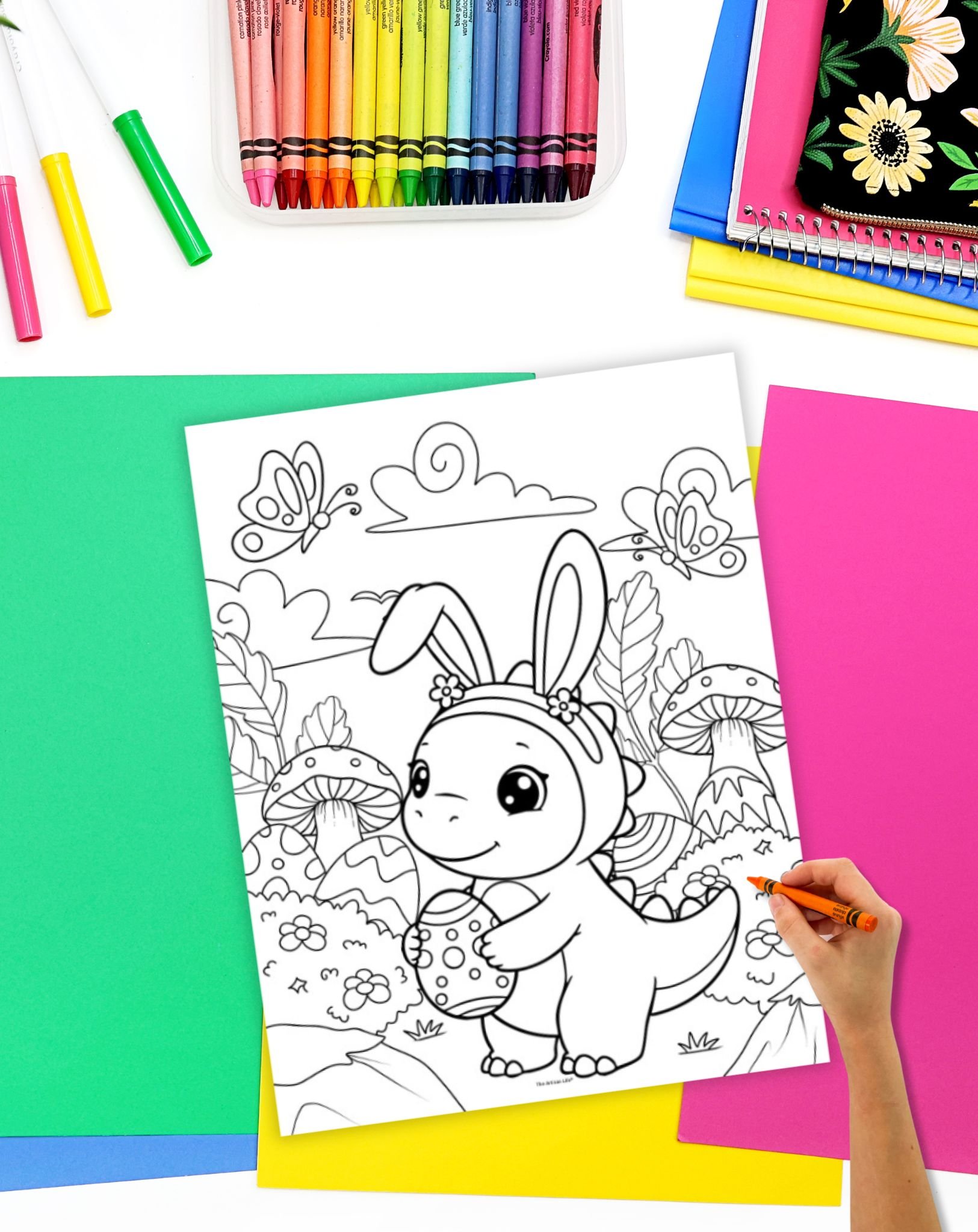 A hand is coloring an Easter-themed coloring page featuring a cartoon dinosaur dressed as a bunny holding an Easter egg among flowers and mushrooms, surrounded by colorful art supplies and paper.
