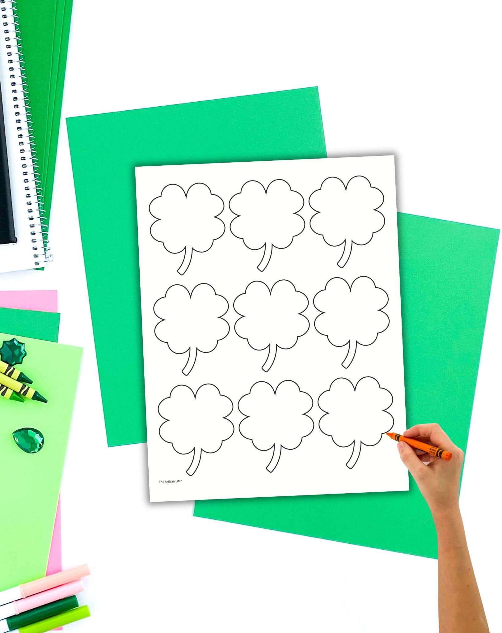 The image features a hand holding an orange crayon, coloring in an outline sheet with nine four leaf clovers, surrounded by various green and pink craft paper and crayons, suitable for St. Patrick’s Day crafts for kids.
