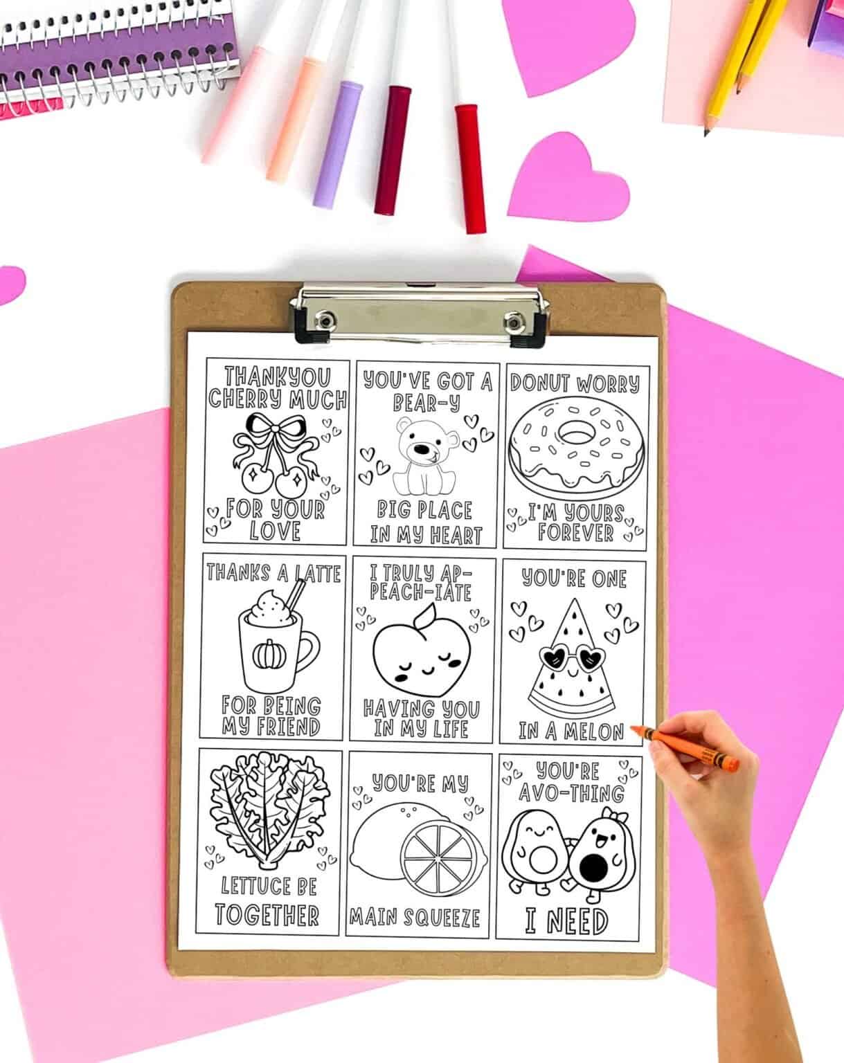 A hand is coloring a printable sheet of Valentine's Day cards featuring humorous food-themed messages, surrounded by colorful markers and decorative paper.