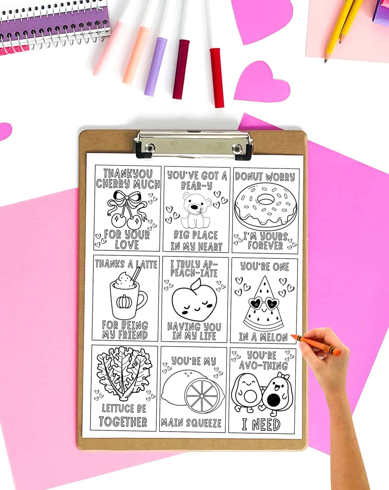 A hand is coloring a printable sheet of Valentine's Day cards featuring humorous food-themed messages, surrounded by colorful markers and decorative paper.
