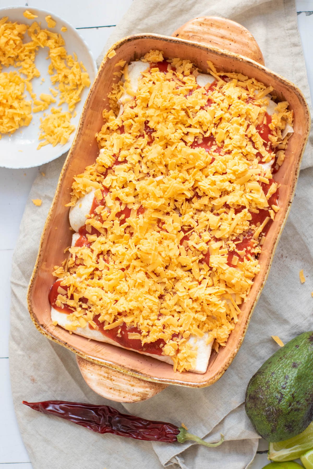 A baking dish filled with rolled black bean enchiladas topped with red sauce, accompanied by shredded cheese and nearby ingredients including an avocado and dried chili peppers.
