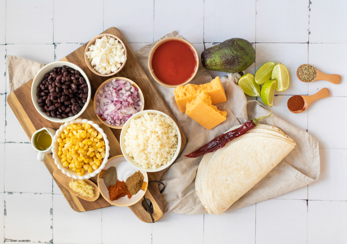 An assortment of ingredients for making black bean enchiladas is displayed on a wooden cutting board, including black beans, corn, cheese, rice, tortillas, red sauce, and various spices, highlighting a healthy, vegetarian meal preparation.
