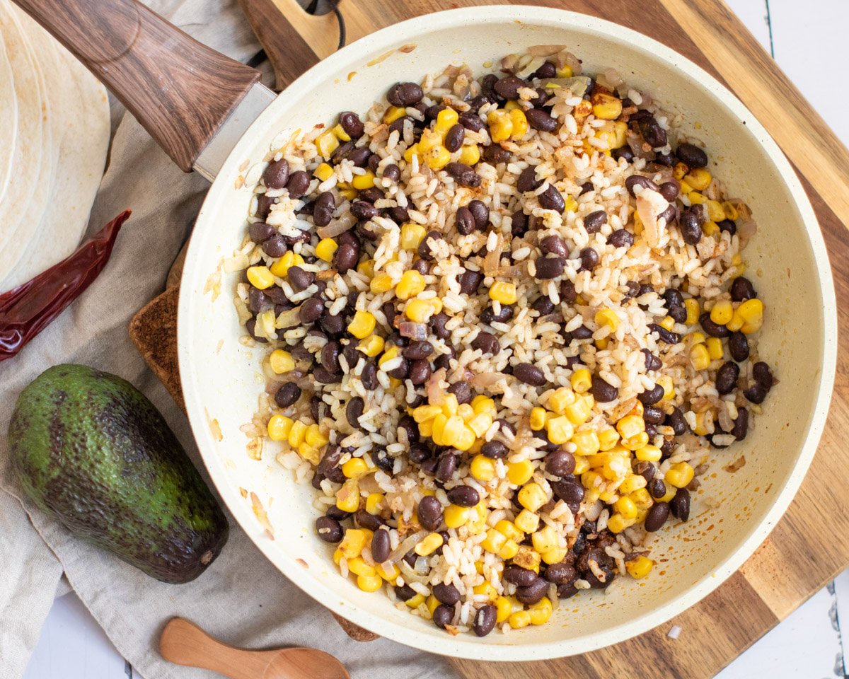 A skillet filled with a mixture of black beans, corn, and rice topped with shredded cheese, prepared as a filling for vegetarian enchiladas.
