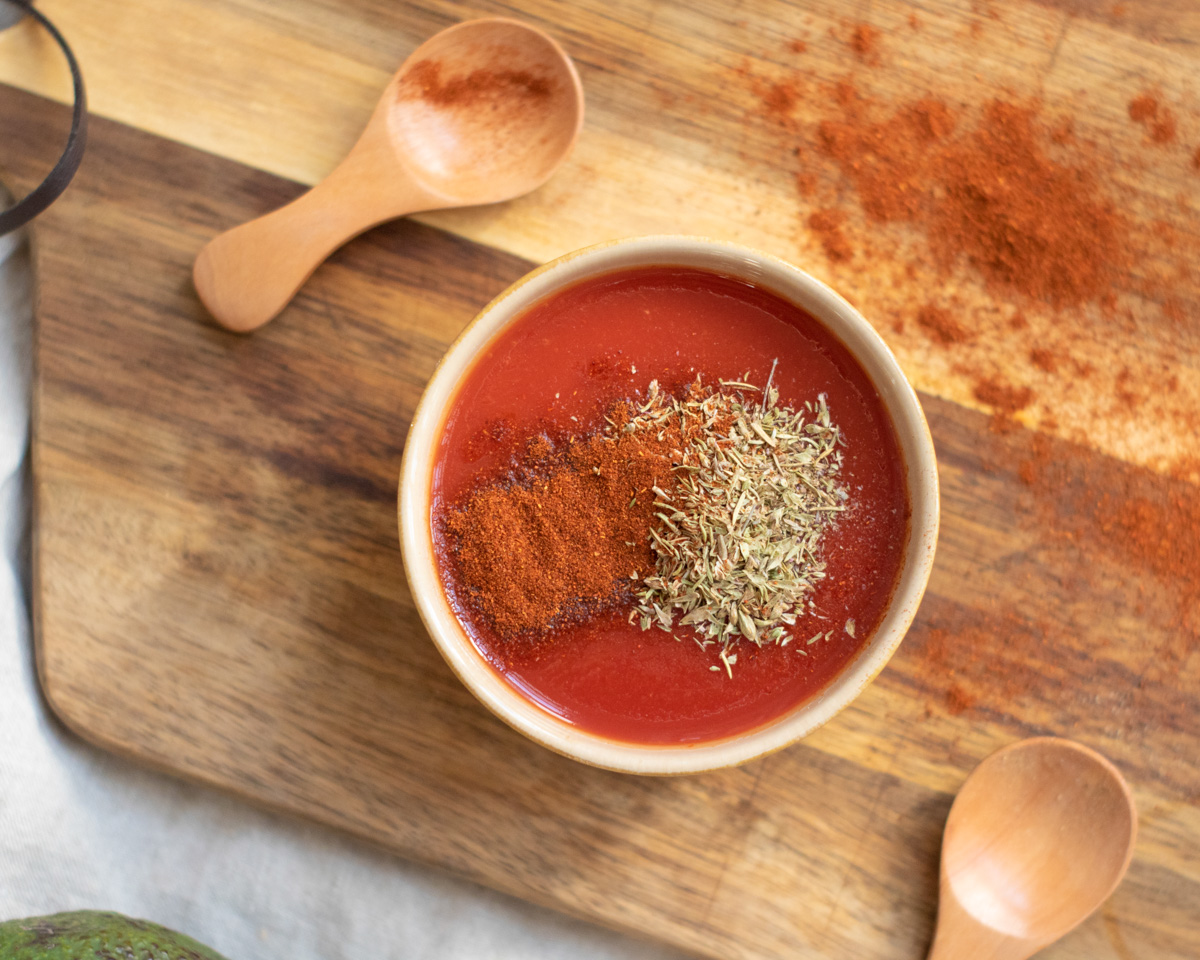 A bowl of red sauce with spices sits on a wooden cutting board beside a small wooden spoon.
