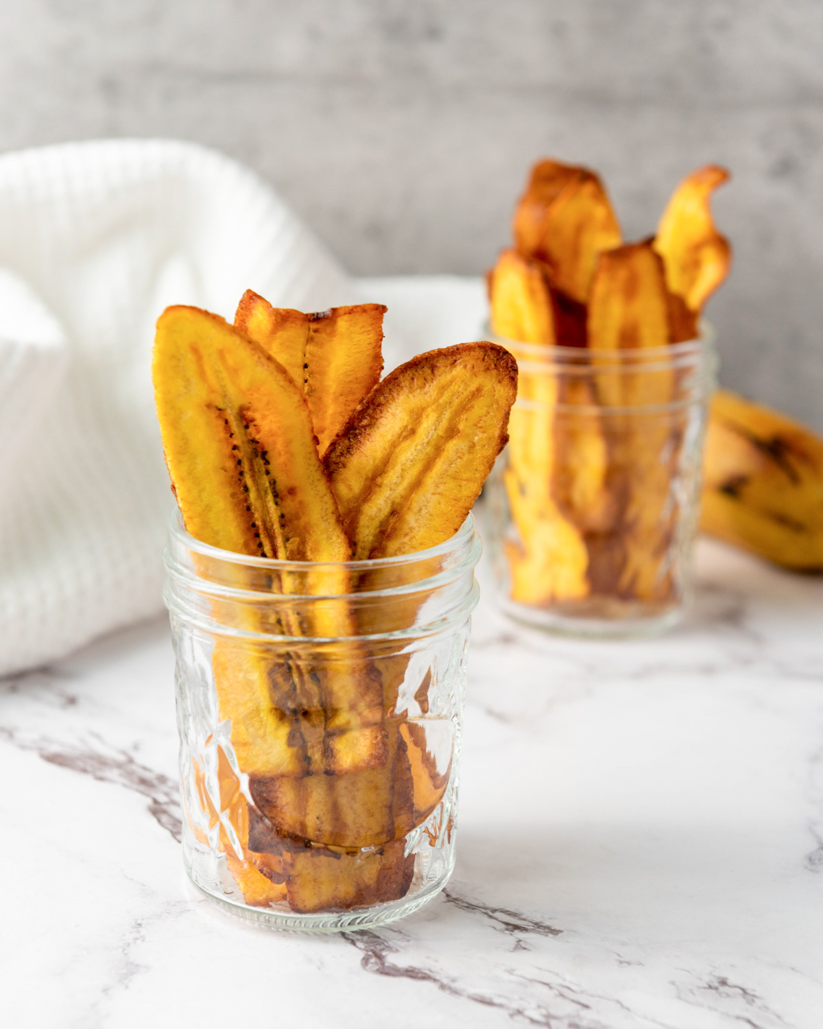 A close-up view of fried plantain chips served in glass jars, with a fresh plantain in the background, showcasing a gluten-free snack option.
