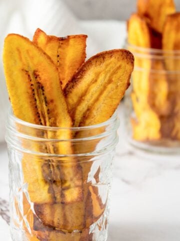 A close-up image of fried plantain chips arranged in glass jars, with a whole ripe plantain positioned in the background, illustrating a simple plantain recipe suitable for gluten-free snacks.