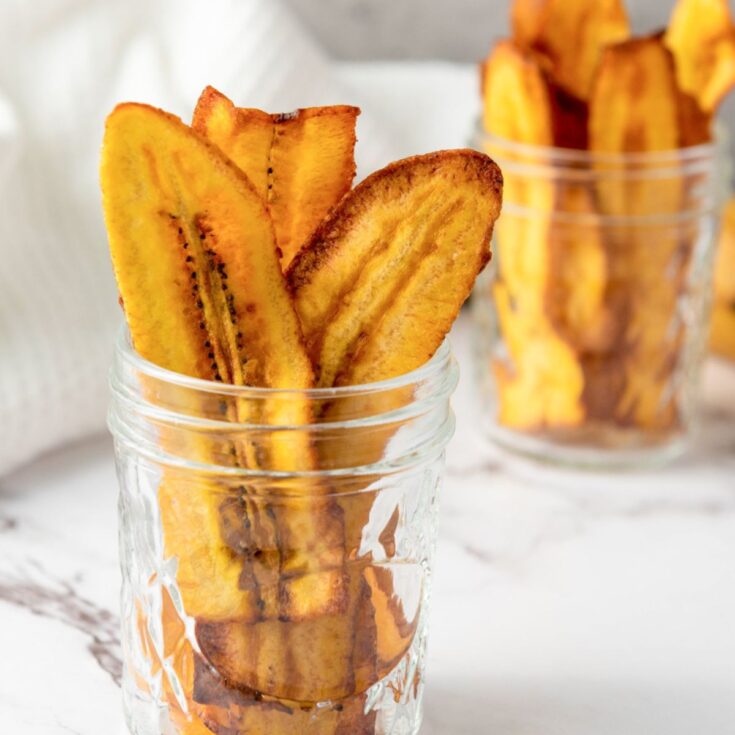 A close-up image of fried plantain chips arranged in glass jars, with a whole ripe plantain positioned in the background, illustrating a simple plantain recipe suitable for gluten-free snacks.