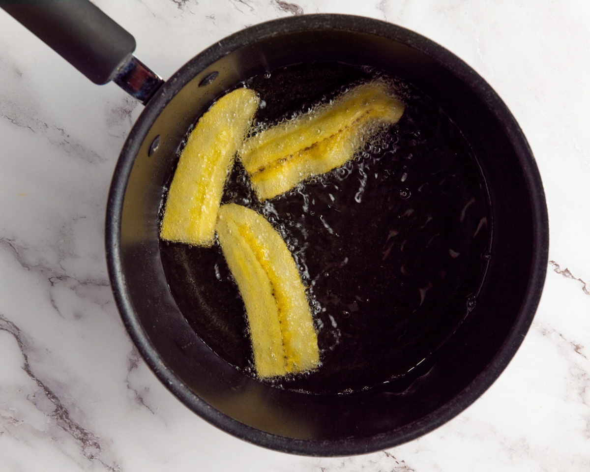 A pot of hot oil contains several frying plantain slices, suggesting preparation for a recipe such as fried plantain chips, common in various cuisines including Mexican and Cuban.
