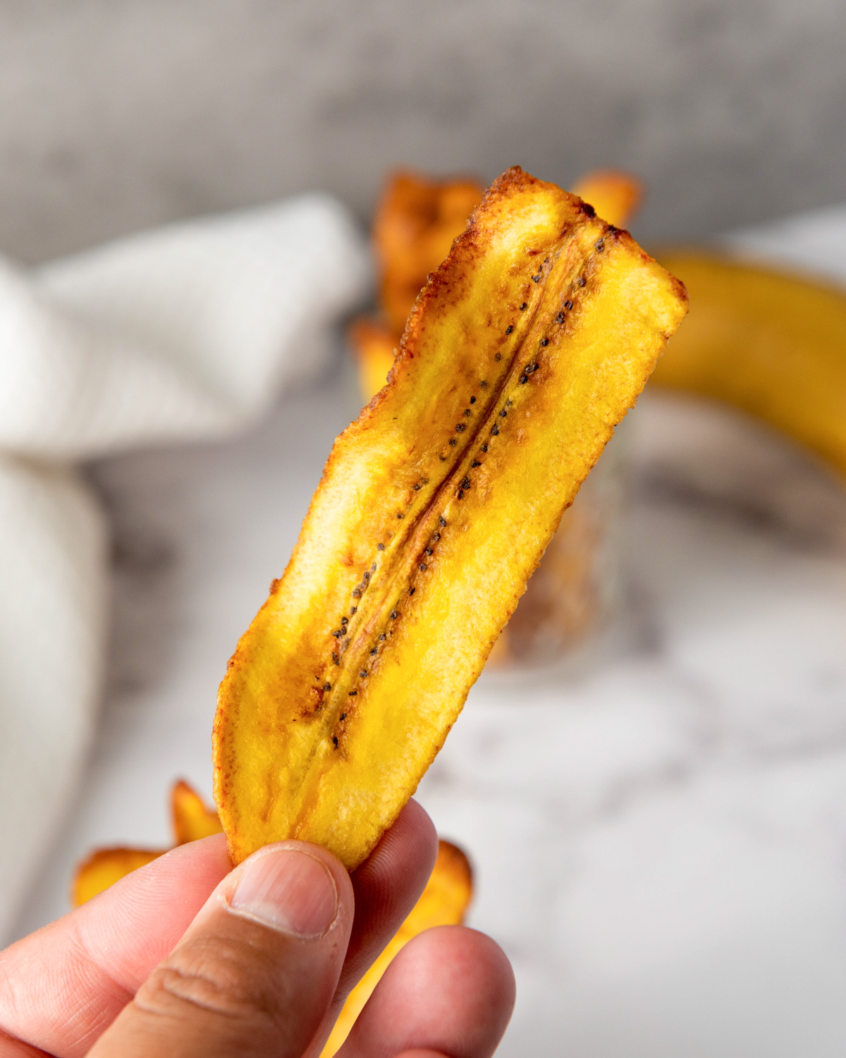 A hand holds a crispy fried plantain chip in front of a jar filled with more chips and a whole plantain in the background, showcasing a gluten-free snack option.

