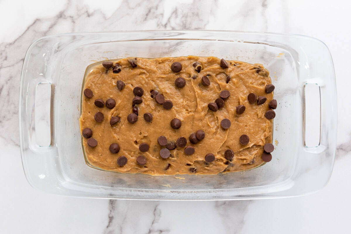 A glass baking dish contains a thick batter for gluten-free banana bread, topped with chocolate chips, set on a marble countertop.
