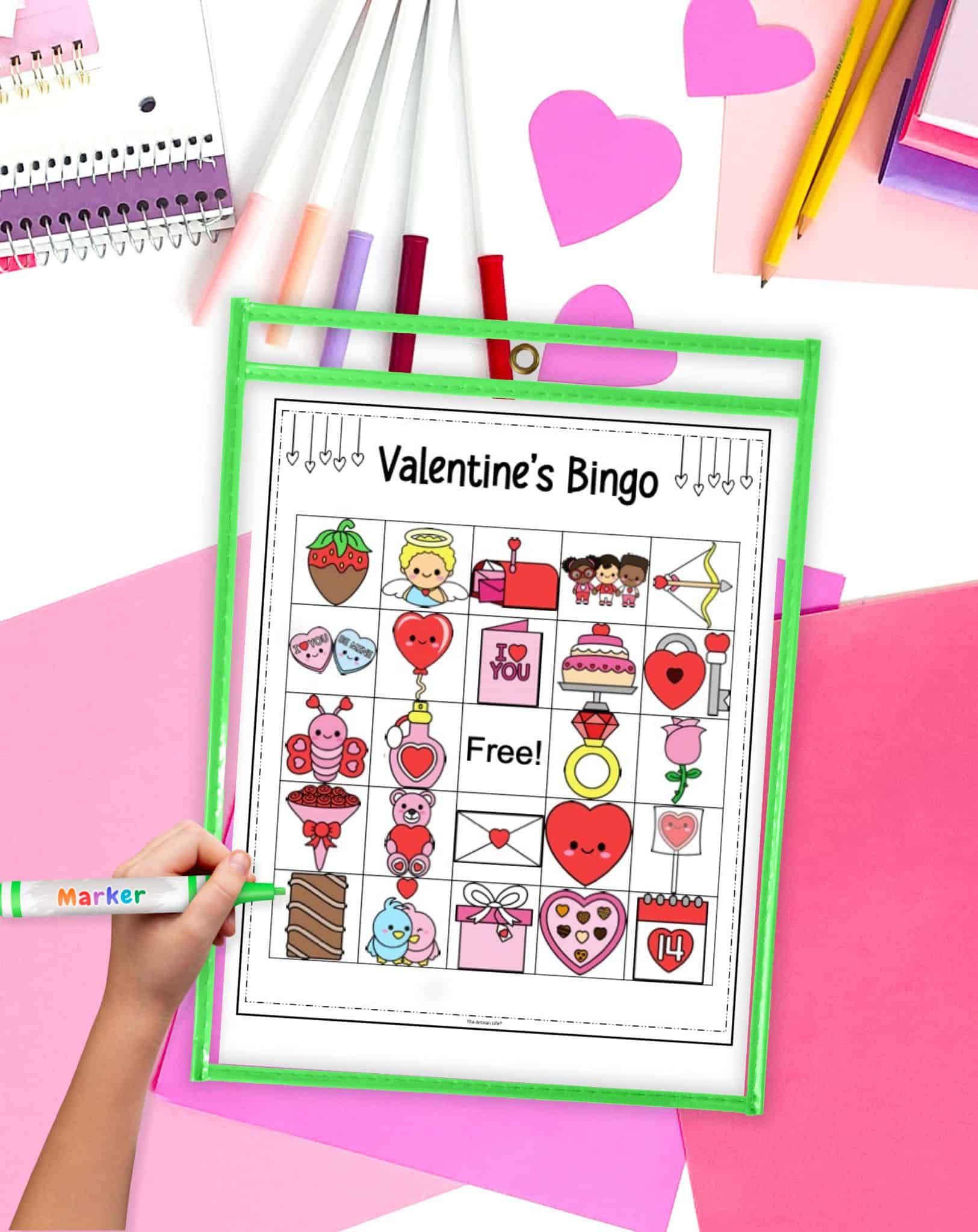 A hand holding a marker is positioned next to a Valentine’s Bingo card displaying various holiday-themed icons, surrounded by colorful stationery and heart-shaped papers, highlighting activities for a Valentine's classroom party or family Valentine’s party.