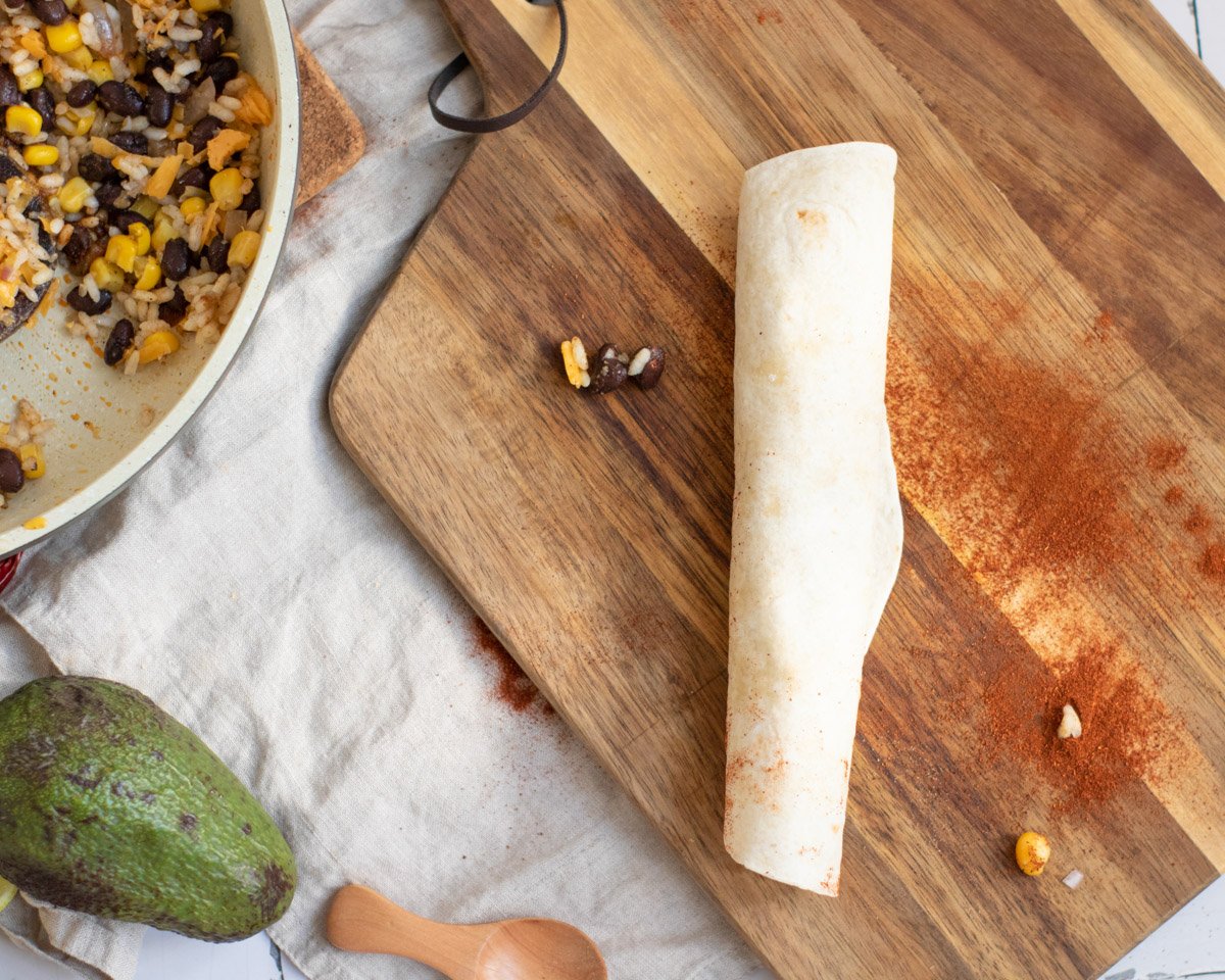 A rolled tortilla filled with a mixture of black beans, corn, and rice sits on a wooden cutting board, alongside an avocado and a spoon, amid spices indicating preparation for black bean enchiladas, a vegetarian dish.
