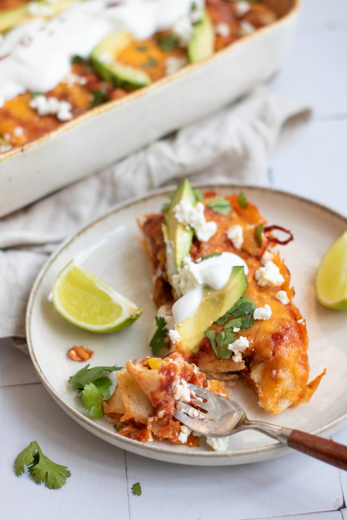 A baked dish of vegetarian enchiladas is topped with avocado slices, sour cream, cilantro, and feta cheese, surrounded by lime wedges on a light surface.
