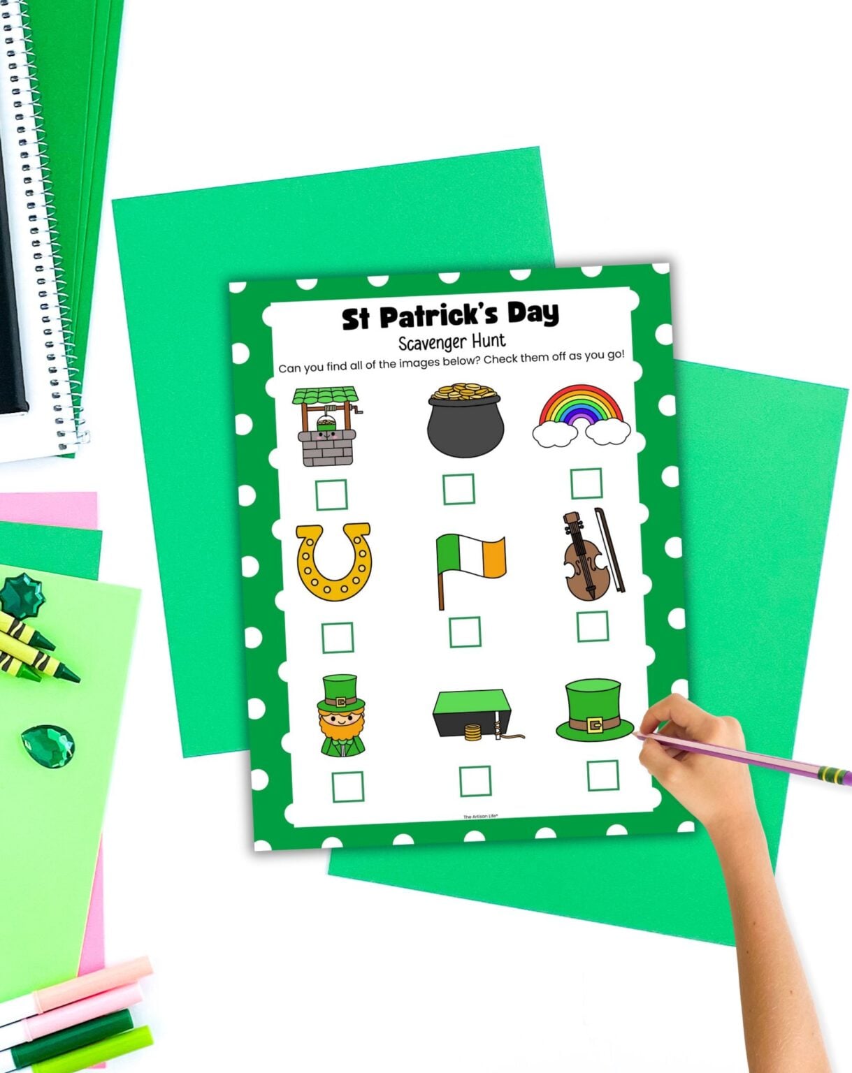 A hand is filling out a St Patrick’s Day scavenger hunt printable sheet, featuring images to find, on a colorful surface with additional green papers and craft supplies around it.