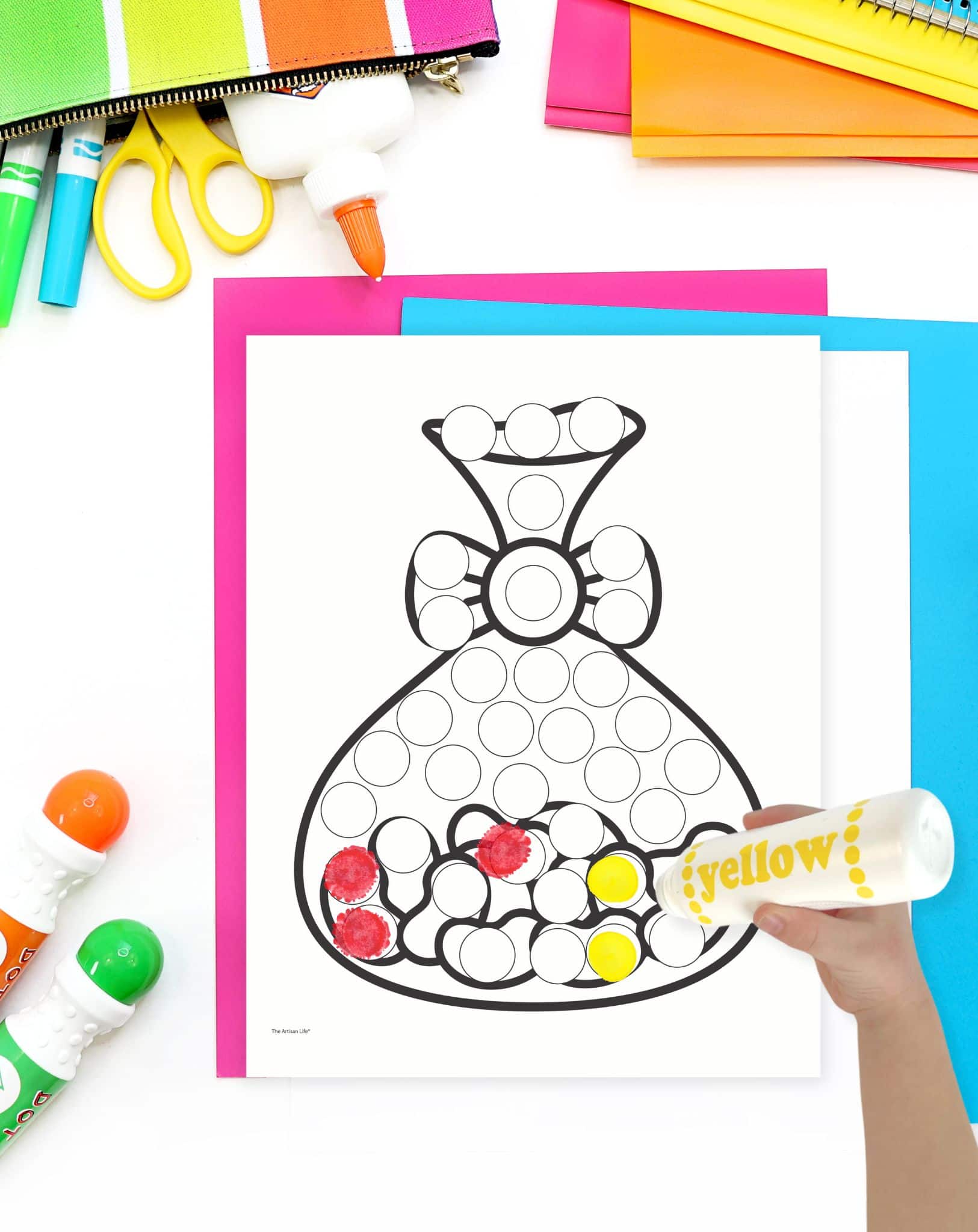 A child's hand is applying yellow dot marker to a dot art printable shaped like a bag, surrounded by colorful craft supplies, highlighting an engaging Easter-themed activity for toddlers.
