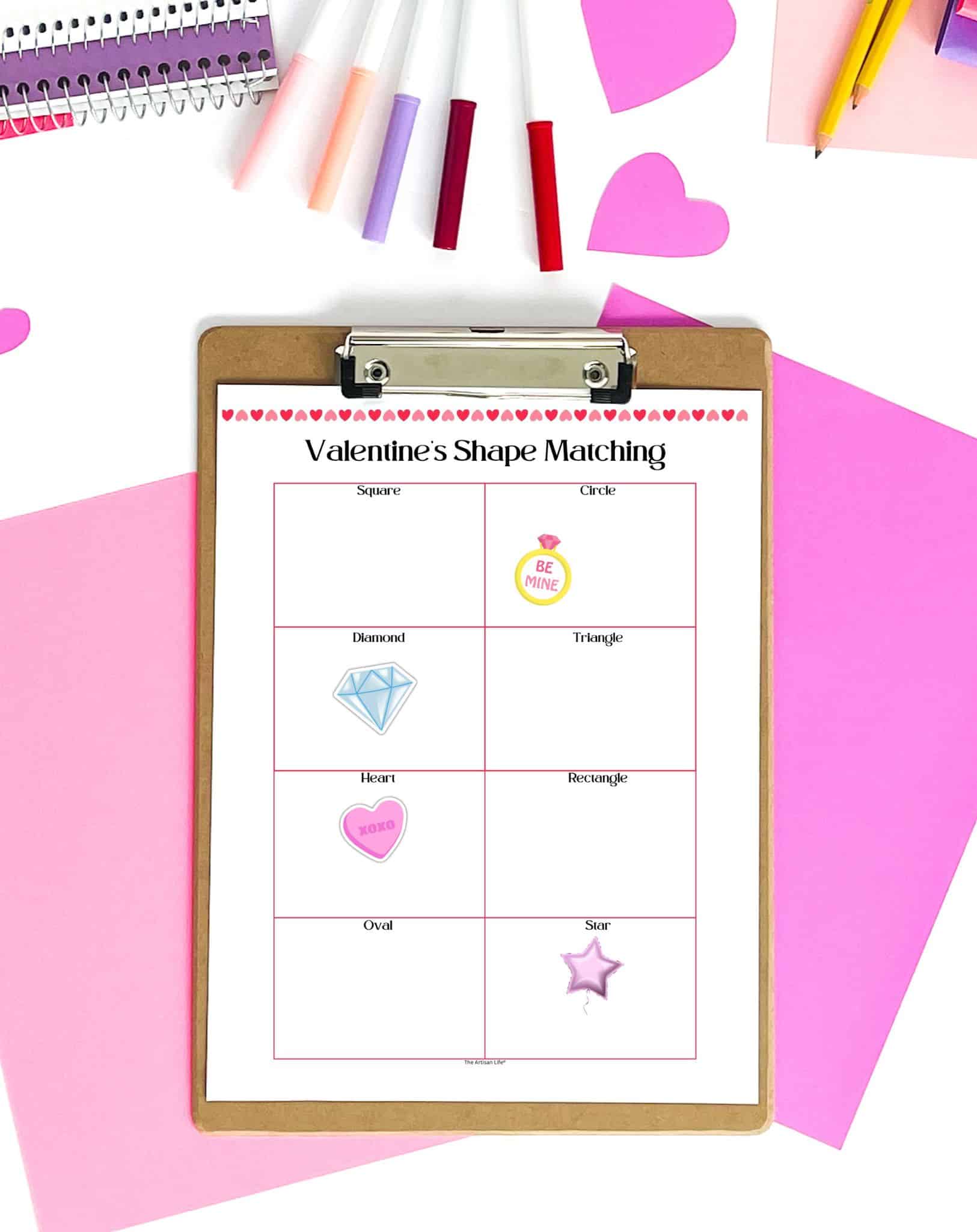 A clipboard displays a "Valentine's Shape Matching" activity sheet featuring various shapes and corresponding images, surrounded by colorful pens and heart cutouts, designed for young children's learning and recognition of shapes. The page is on a clip board