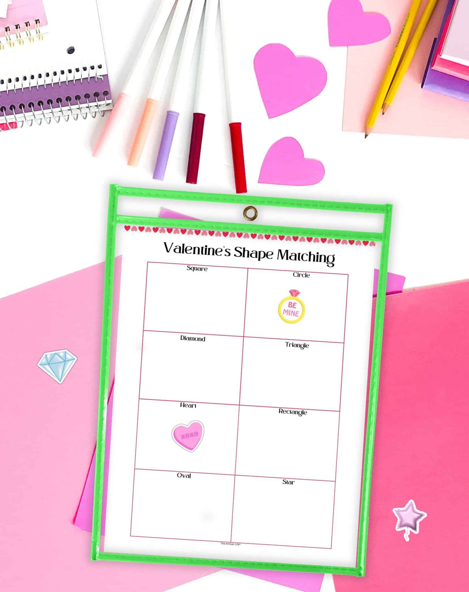 A clipboard displays a "Valentine's Shape Matching" activity sheet featuring various shapes and corresponding images, surrounded by colorful pens and heart cutouts, designed for young children's learning and recognition of shapes.
