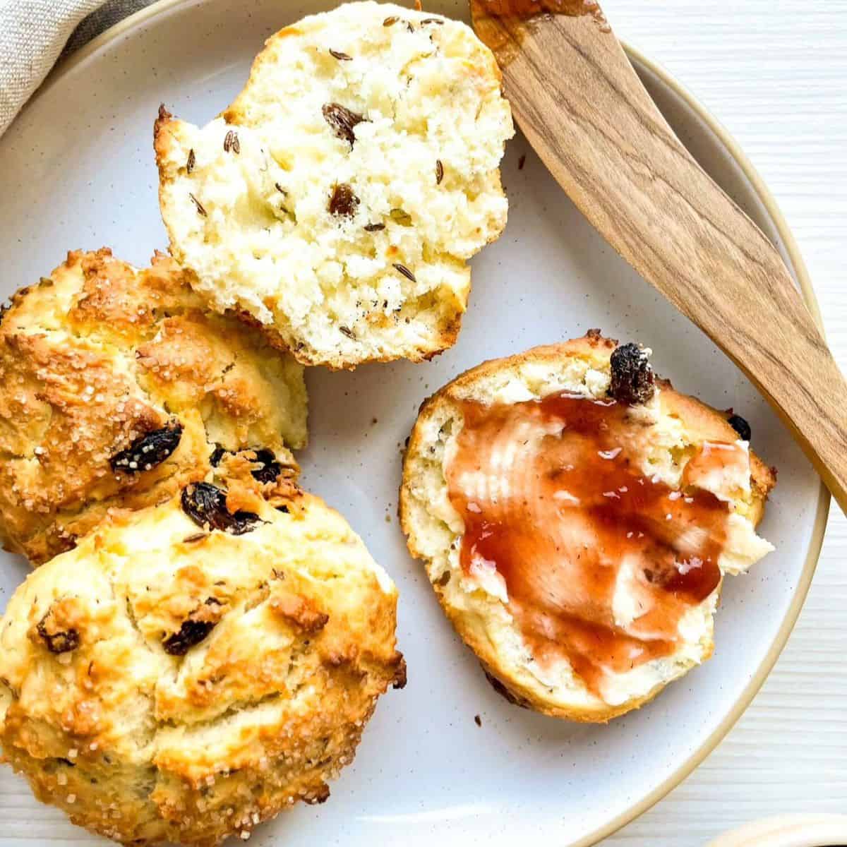 A plate of freshly baked Irish soda bread scones, some cut open to reveal a soft interior with raisins, accompanied by a wooden butter knife and a small bowl of raisins.
