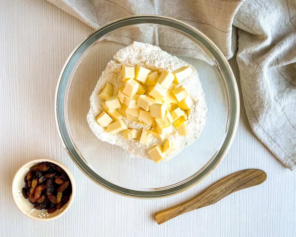 A glass bowl containing flour and cubed butter is placed on a table alongside a small bowl of raisins and a wooden spatula, suggesting preparation for making buttermilk scones or Irish soda bread scones.
