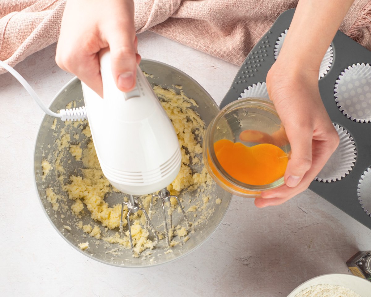 A person is shown adding egg to a bowl of mixed cake batter while preparing to make cupcakes, with cupcake liners ready in a tray nearby.
