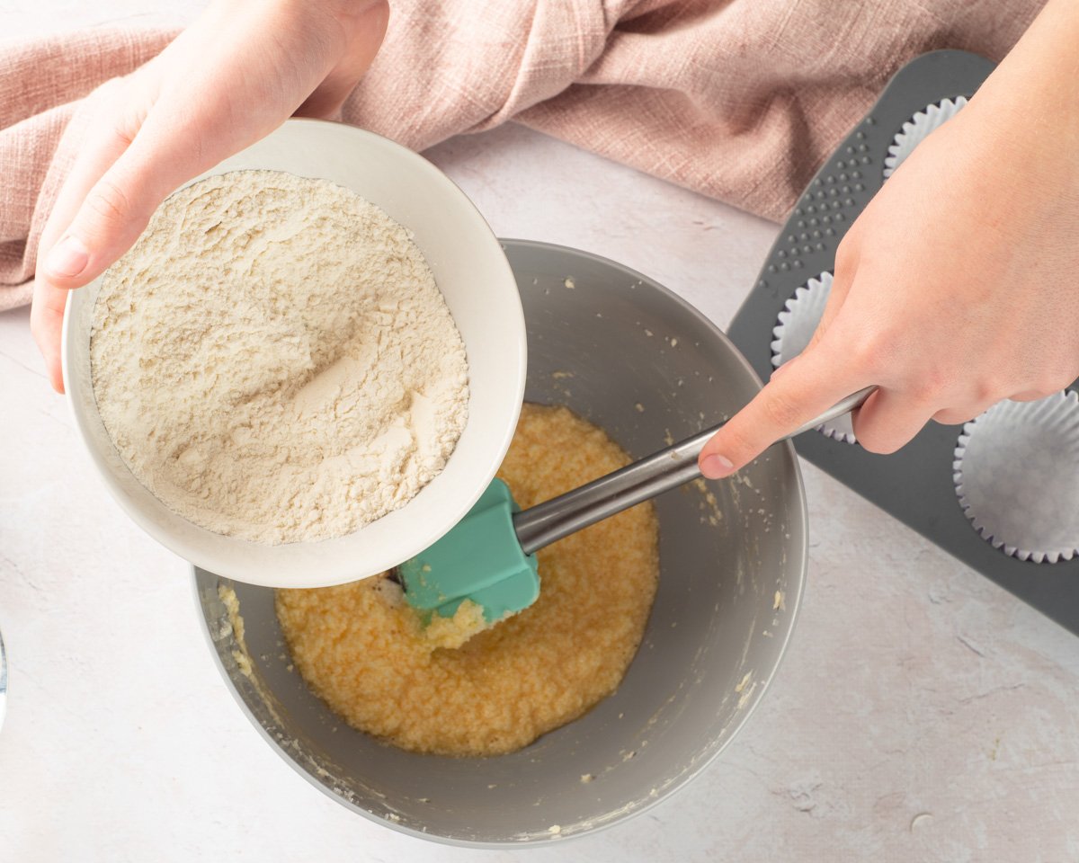 A person is shown adding gluten-free flour to a bowl of mixed cake batter while preparing to make cupcakes, with cupcake liners ready in a tray nearby.
