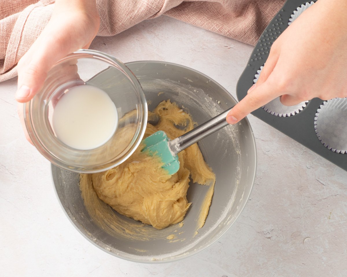 A person is shown adding milk to a bowl of mixed cake batter while preparing to make cupcakes, with cupcake liners ready in a tray nearby.
