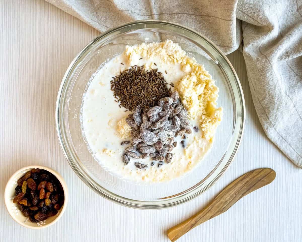 A glass bowl contains a mixture of wet ingredients and dry ingredients, including buttermilk, flour, and raisins, with a small bowl of mixed raisins on the side, set on a light wooden surface beside a linen cloth and wooden spoon, suggesting preparation for Irish soda bread or scones.
