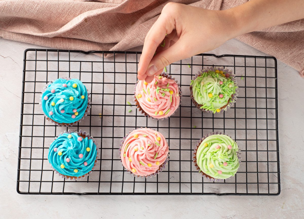 A hand sprinkles colorful toppings on frosted gluten-free vanilla cupcakes arranged on a cooling rack, showcasing ideas for easy Easter desserts.
