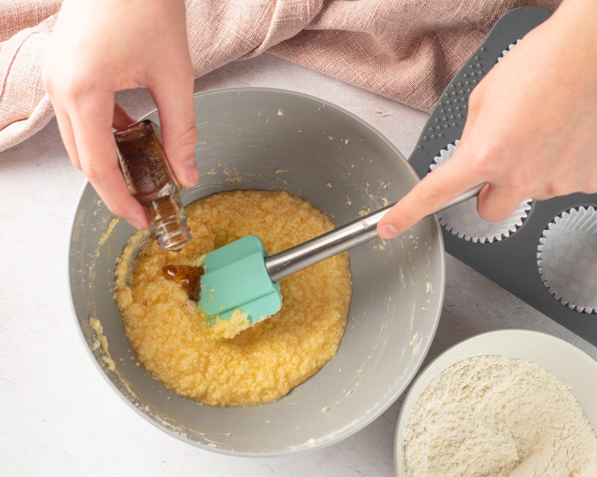 A person is shown adding vanilla to a bowl of mixed cake batter while preparing to make cupcakes, with cupcake liners ready in a tray nearby.
