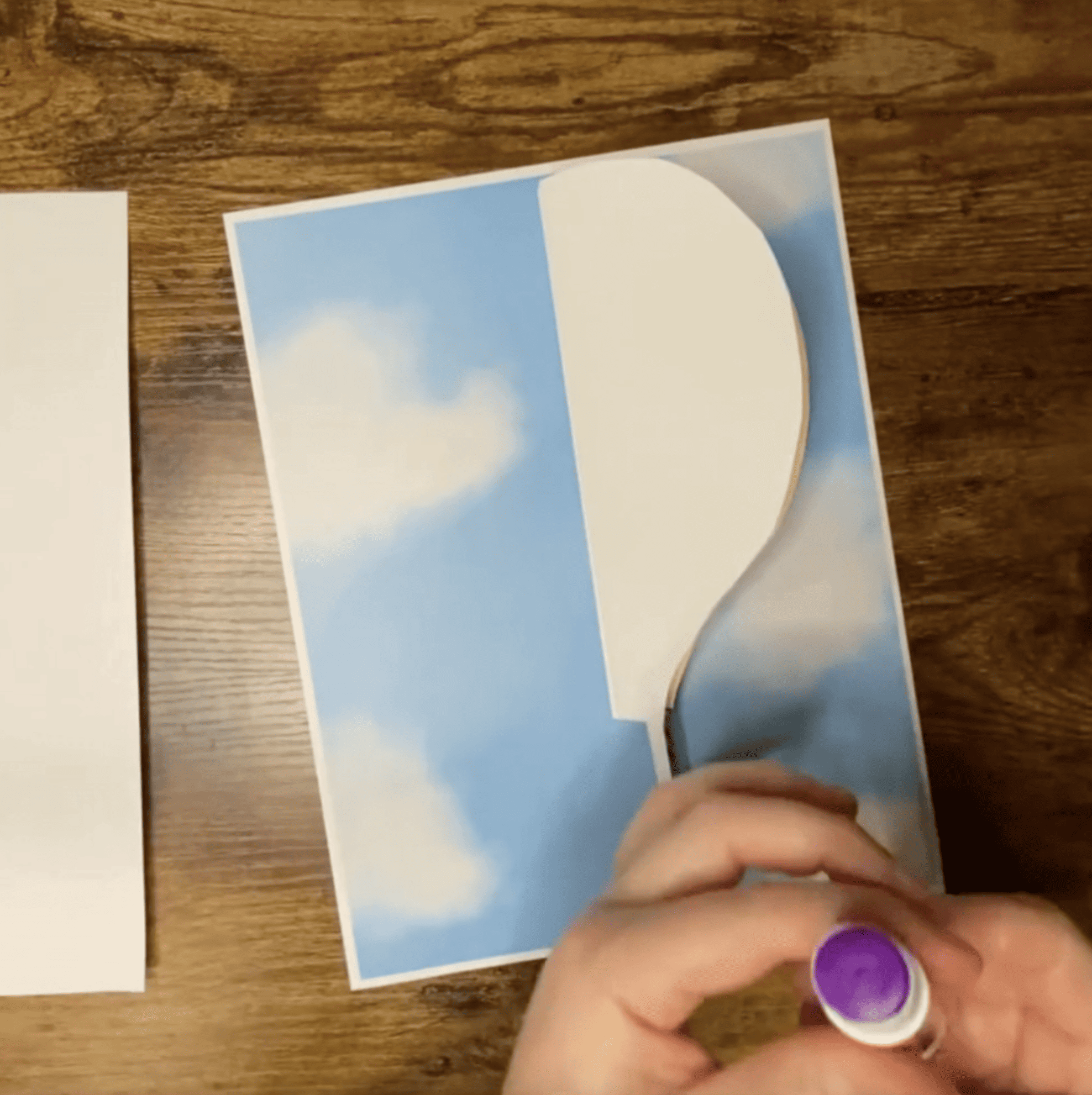 A hand holds a purple glue stick while preparing to assemble a paper hot air balloon craft on a blue sky background, with additional blank paper nearby.
